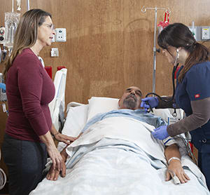 Health care provider caring for man in intensive care unit bed while woman stands nearby.