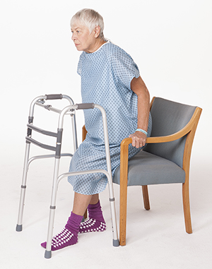 Woman in hospital gown sitting down in chair with walker in front of her.