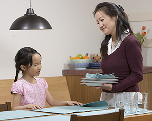 Girl helping woman set dinner table.